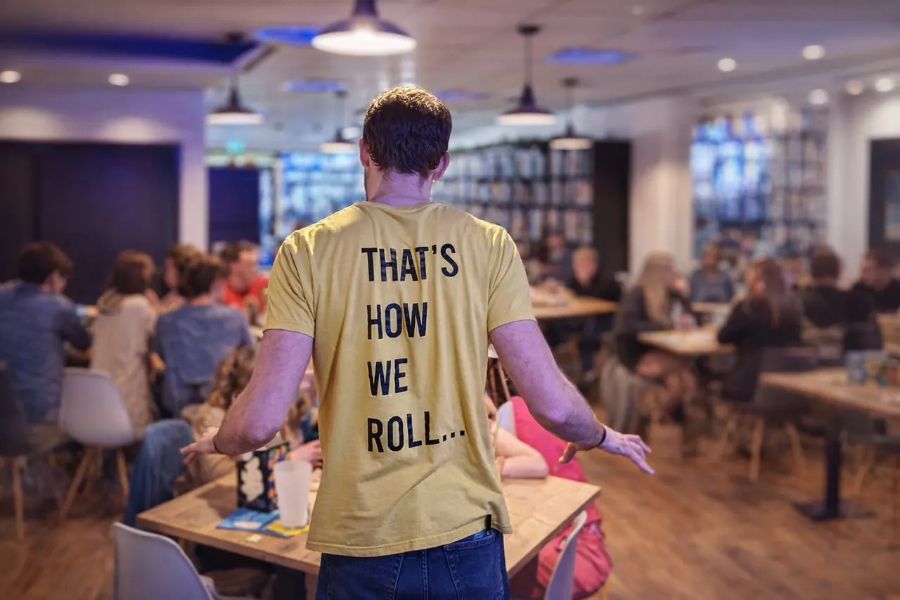 Man in "That's How We Roll" t-shirt stands at bowling alley with other players seated at tables behind him.