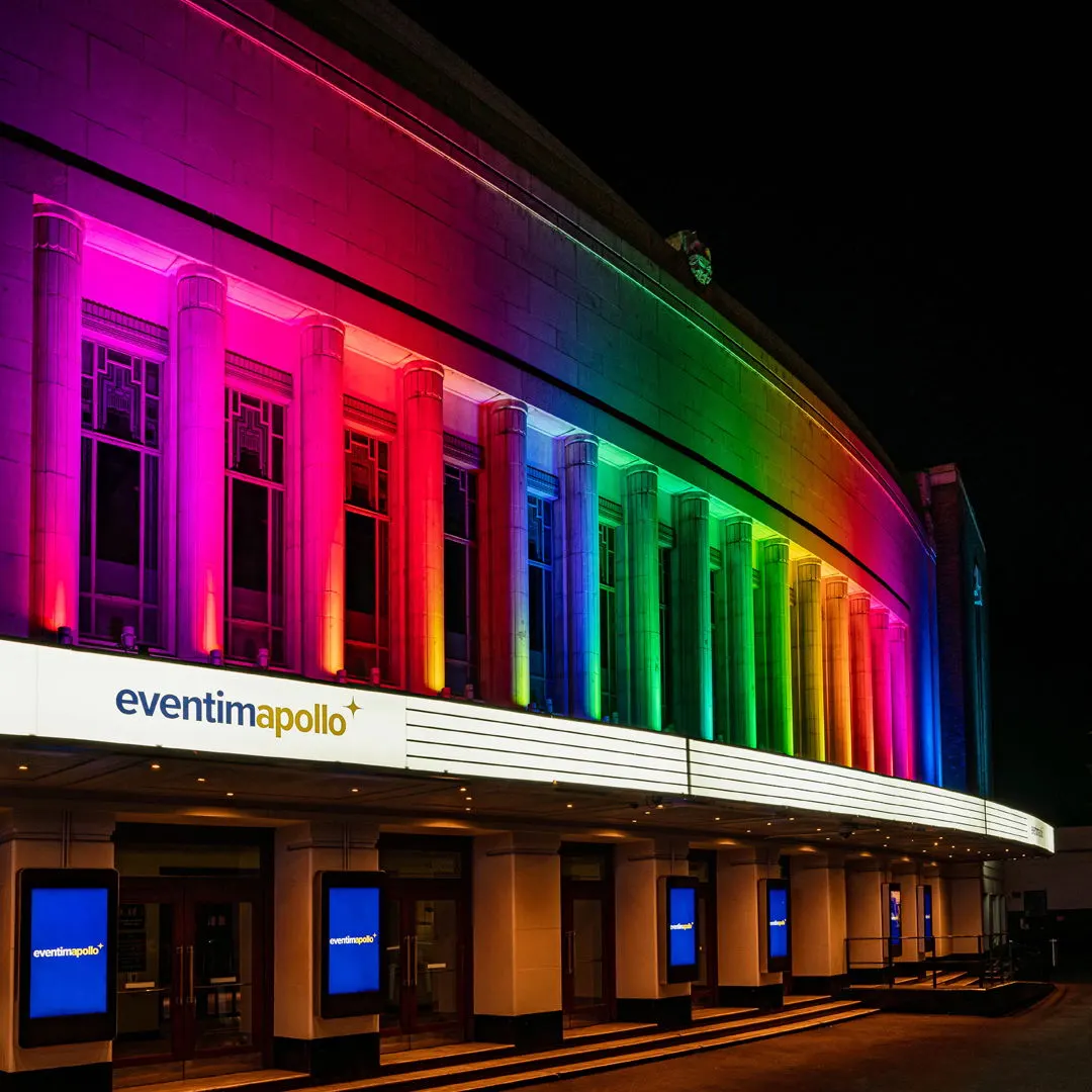 Historic theatre building illuminated at night with rainbow-coloured neon lighting across its classical facade.