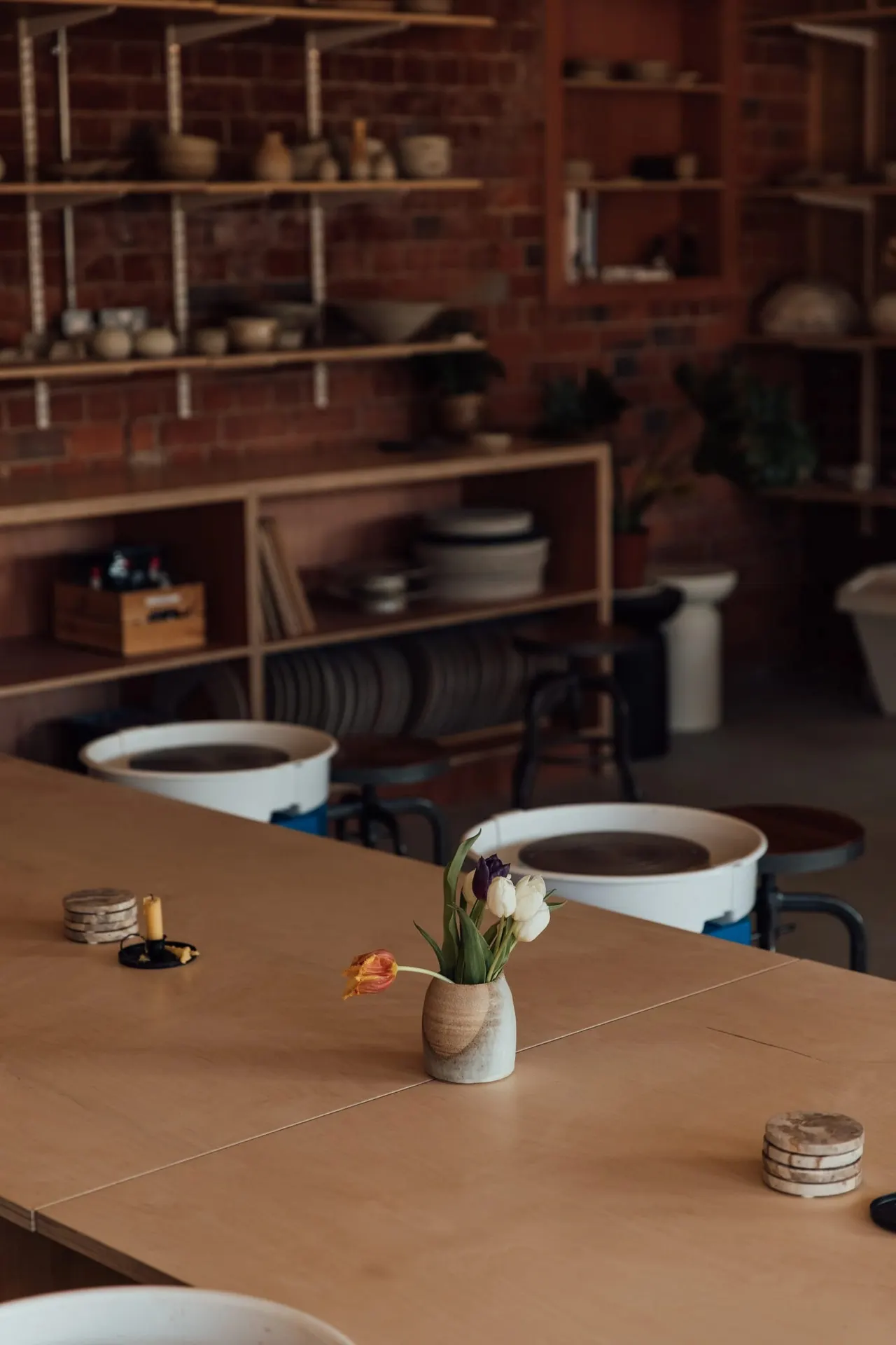Two white mugs of coffee sit on a wooden table with a small vase of white and orange flowers, in a pottery studio with shelving displaying ceramic pieces.