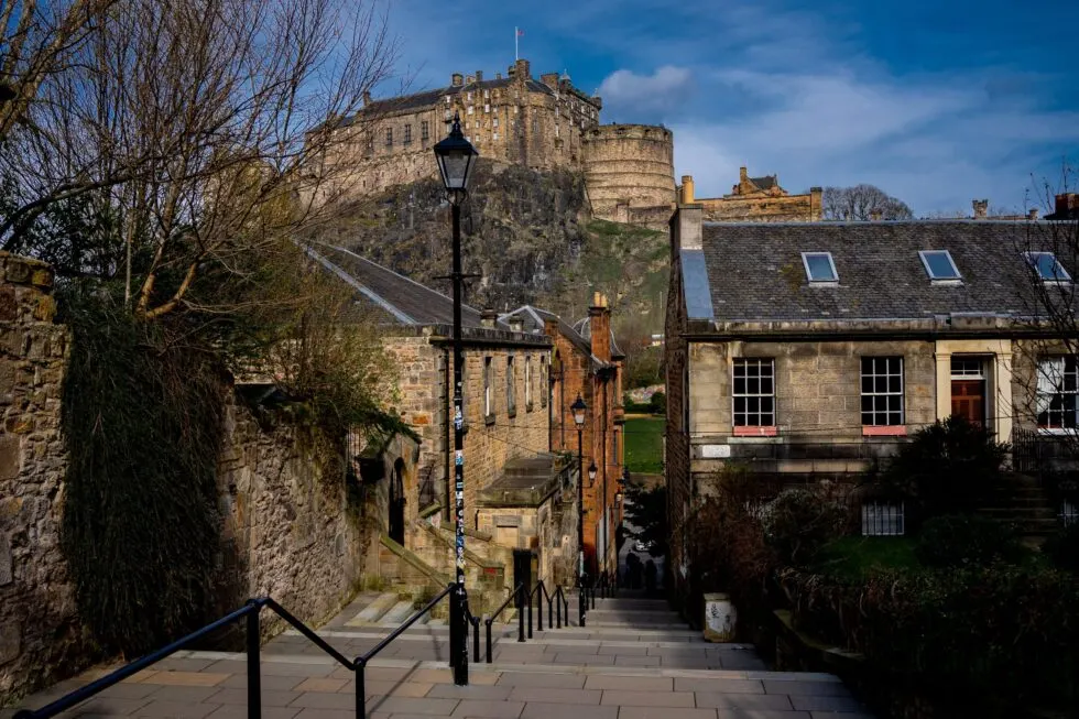A narrow historic alleyway with stone buildings and steps leading towards Edinburgh Castle on the hilltop.
