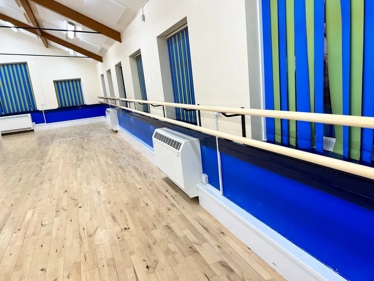 Empty dance studio with wooden sprung floor, blue and green striped curtains, ballet barre, and white heating units.