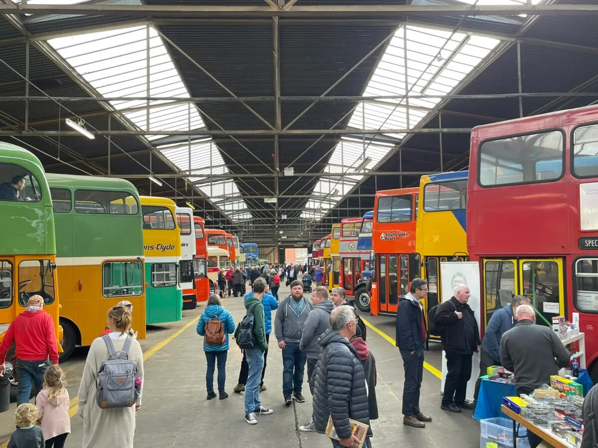Visitors exploring a collection of colourful vintage buses and coaches in a covered hangar with steel framework and skylights.