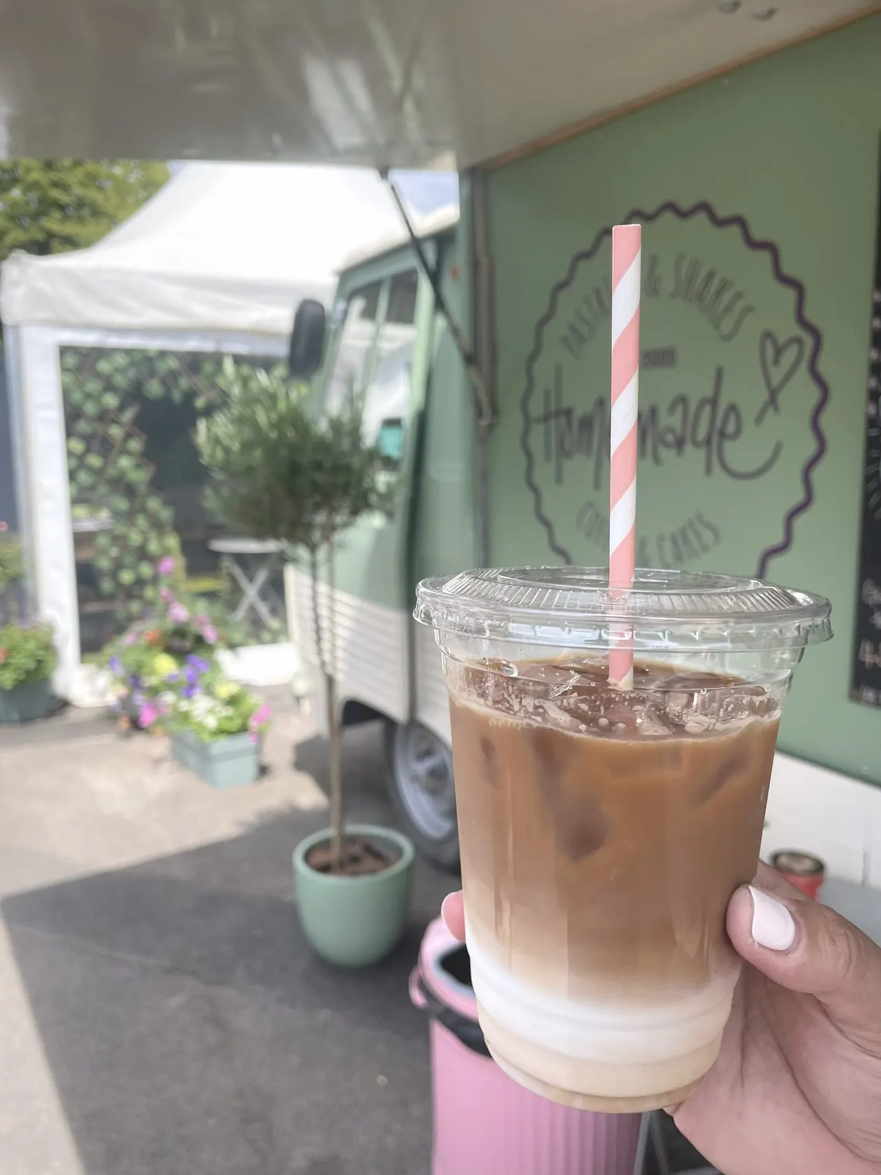 Hand holding an iced coffee drink with striped straw at a café outdoor seating area.