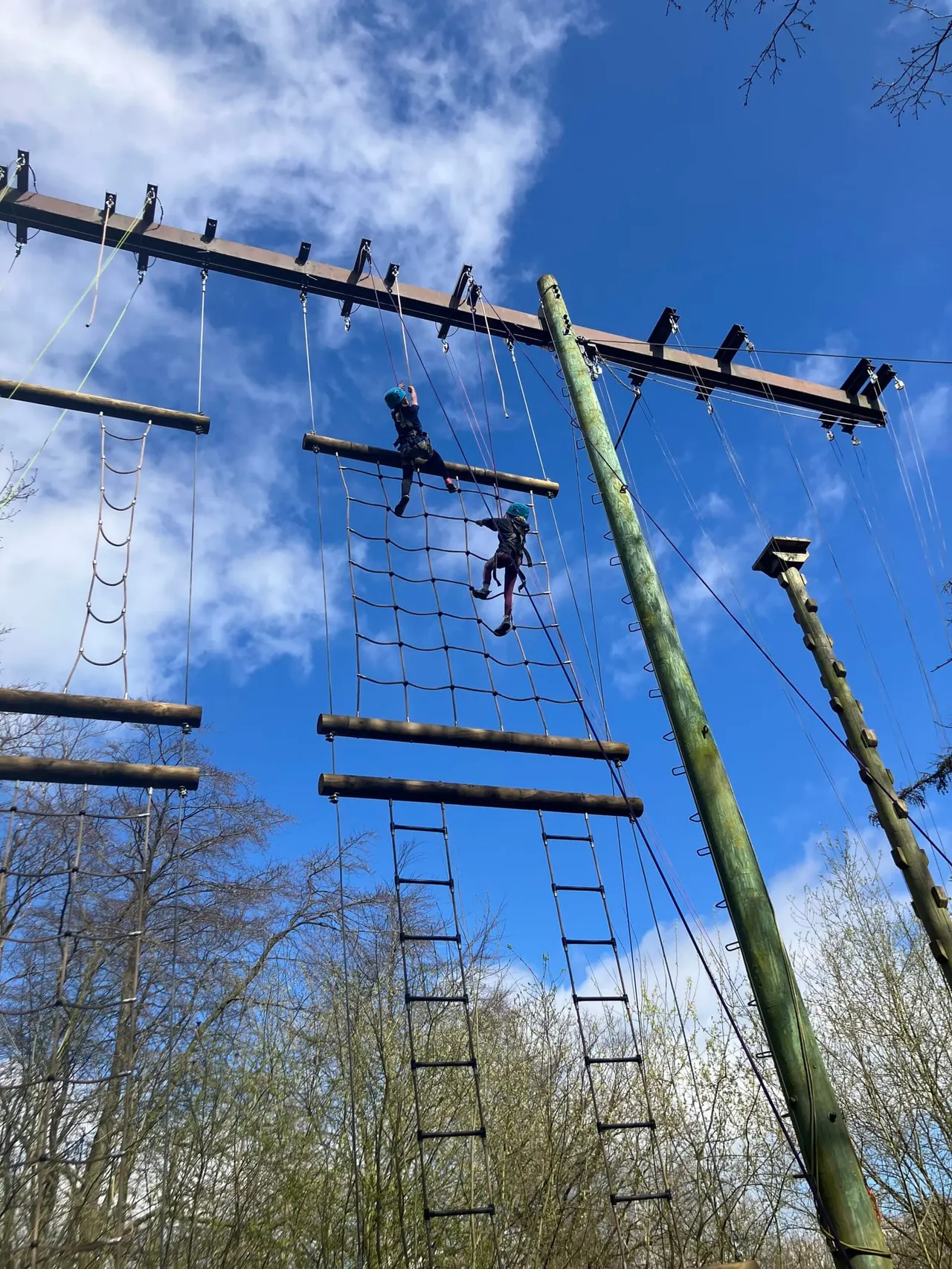 Two people climb a rope net obstacle course whilst others wait on platforms above under a clear blue sky.