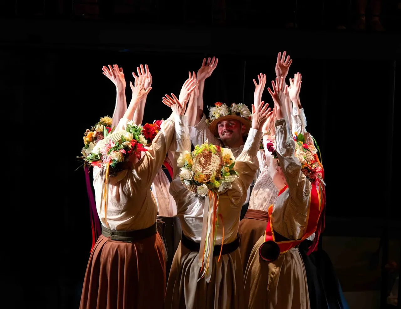 Theatrical performers in colourful costumes with large floral arrangements raise their arms in celebration on stage against a black backdrop.