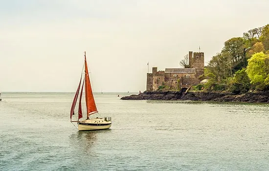 Sailing boat with red sails on calm water near a medieval castle on the shore.