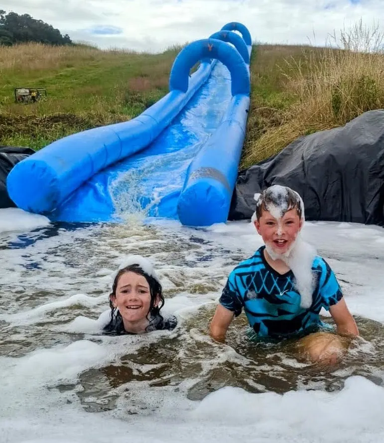 Two children playing in muddy water with a blue inflatable water slide in a rural field.