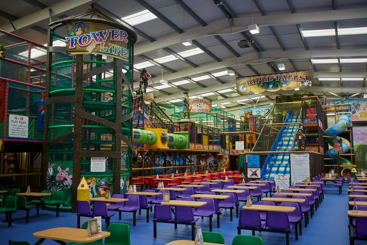 Indoor play centre with colourful climbing structures, slides and rides, with seating areas and tables in the foreground.