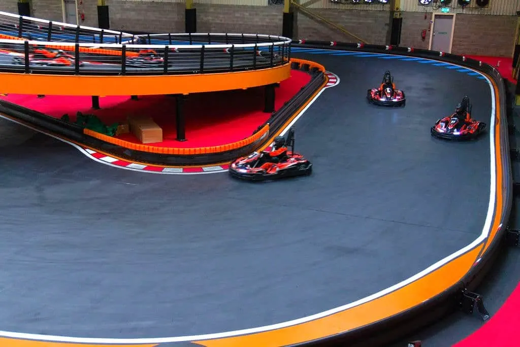 Three go-karts racing on an indoor track with orange and blue barriers inside a brick-walled facility.