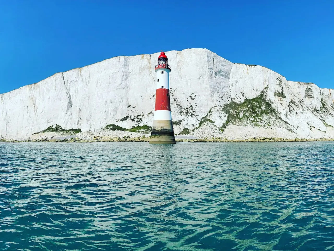 Red and white striped lighthouse standing in sea against white chalk cliffs under clear blue sky.