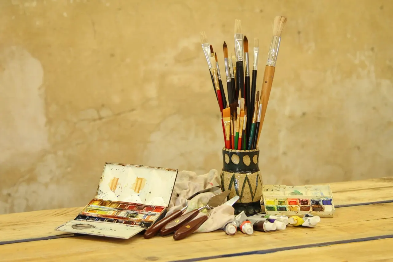 Collection of paintbrushes and art supplies arranged on a wooden table against a textured wall.
