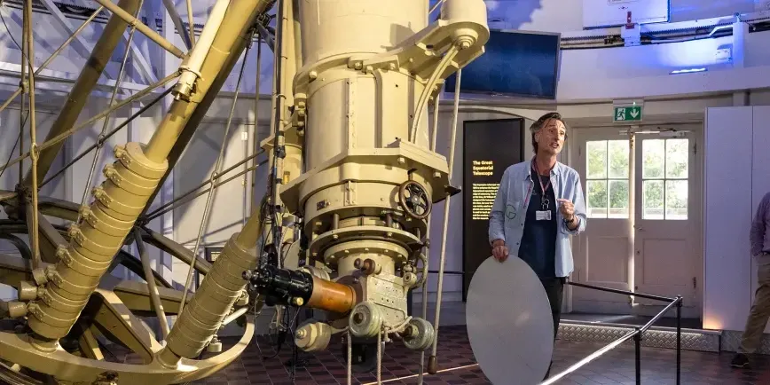 An adult presents to a large brass telescope mechanism in an indoor museum gallery space.