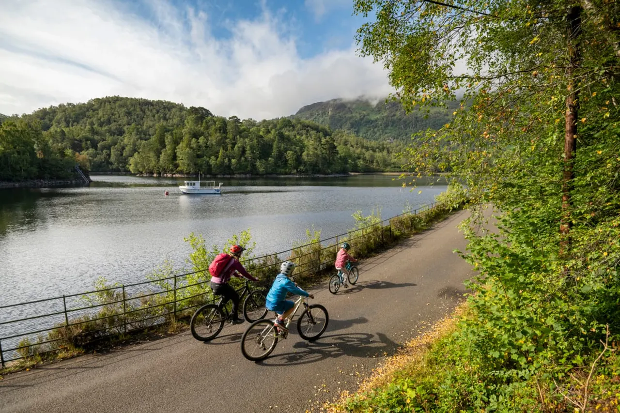 Adults and children cycling on a lakeside path beside forested mountains and a moored boat on the water.
