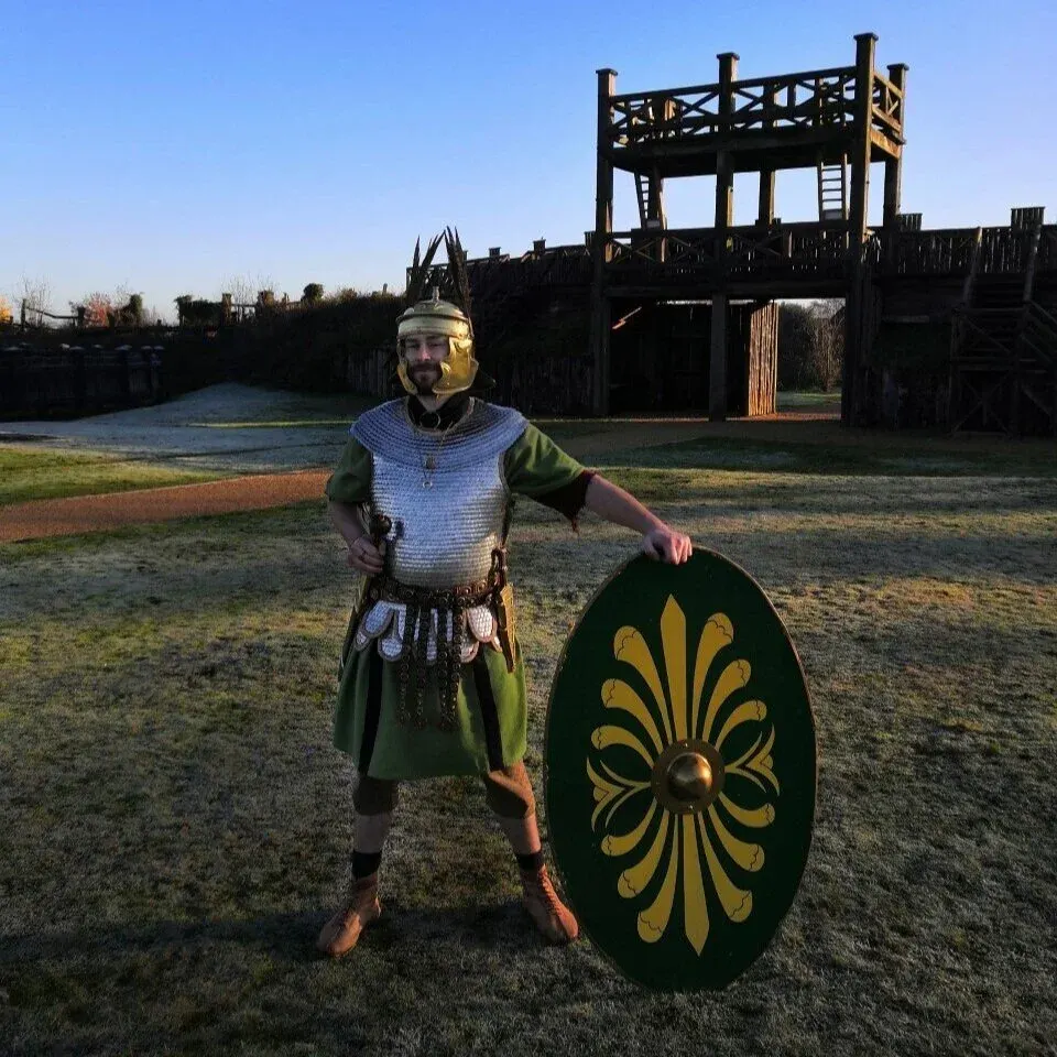 A man in Romano-British military costume holds a decorated shield in front of reconstructed wooden fort buildings.