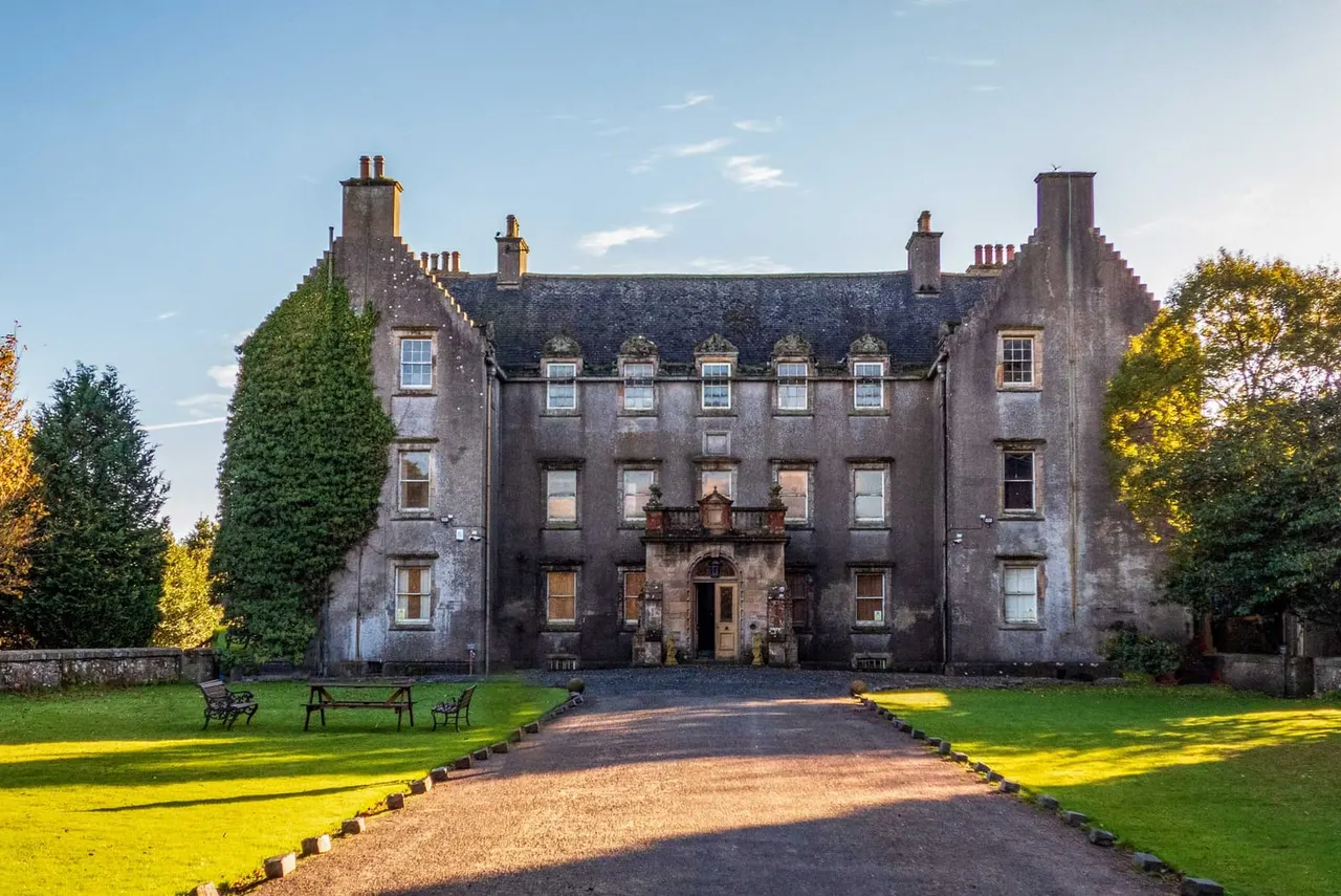 Historic stone mansion with symmetrical facade, multiple chimneys, and manicured gardens with benches, approached via gravel driveway.