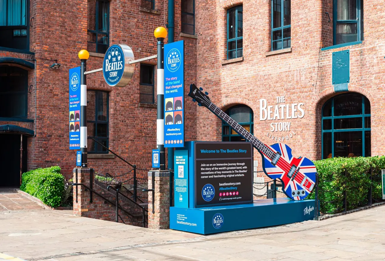 Exterior of The Beatles Story museum in Liverpool, featuring a red brick building with blue signage, a giant guitar sculpture painted with the Union Jack flag, and promotional banners.