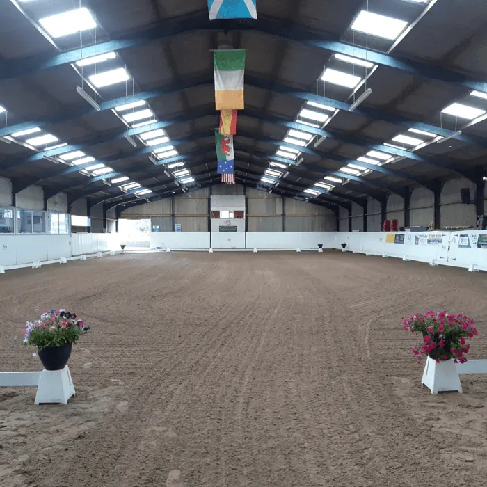 Empty indoor equestrian arena with sand surface, decorative flower planters and white railings.