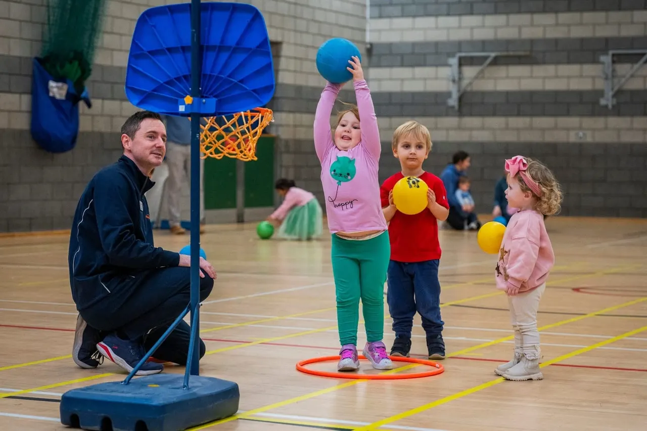 An adult instructor supervises young children playing with balls and hoops during a sports activity in an indoor gymnasium.