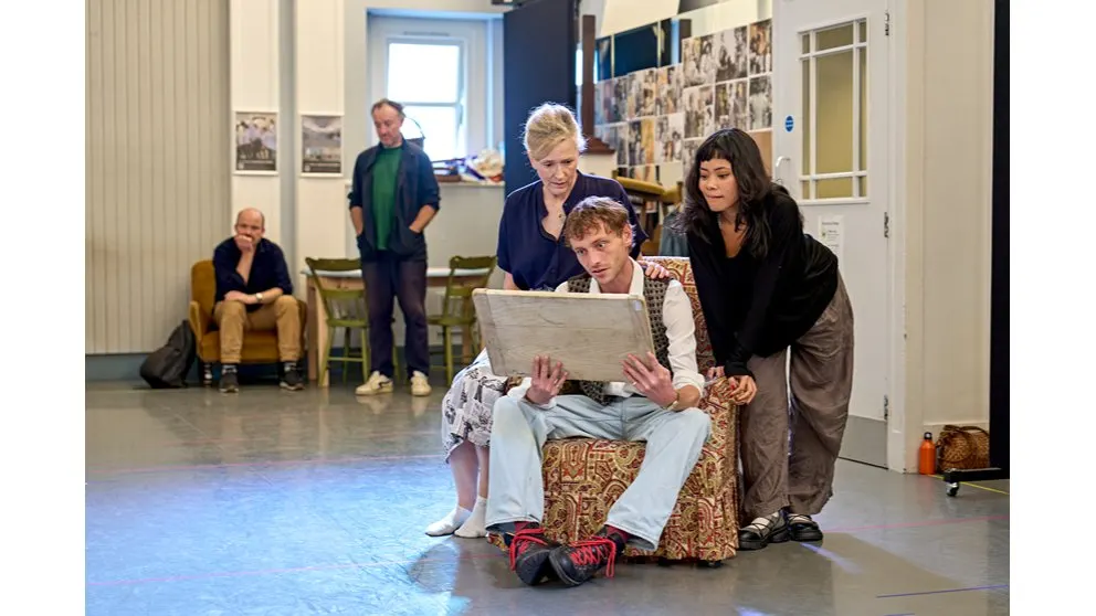 A young person sits in a patterned armchair holding a laptop whilst two women stand beside them during a theatre rehearsal, with other cast members visible in the background.