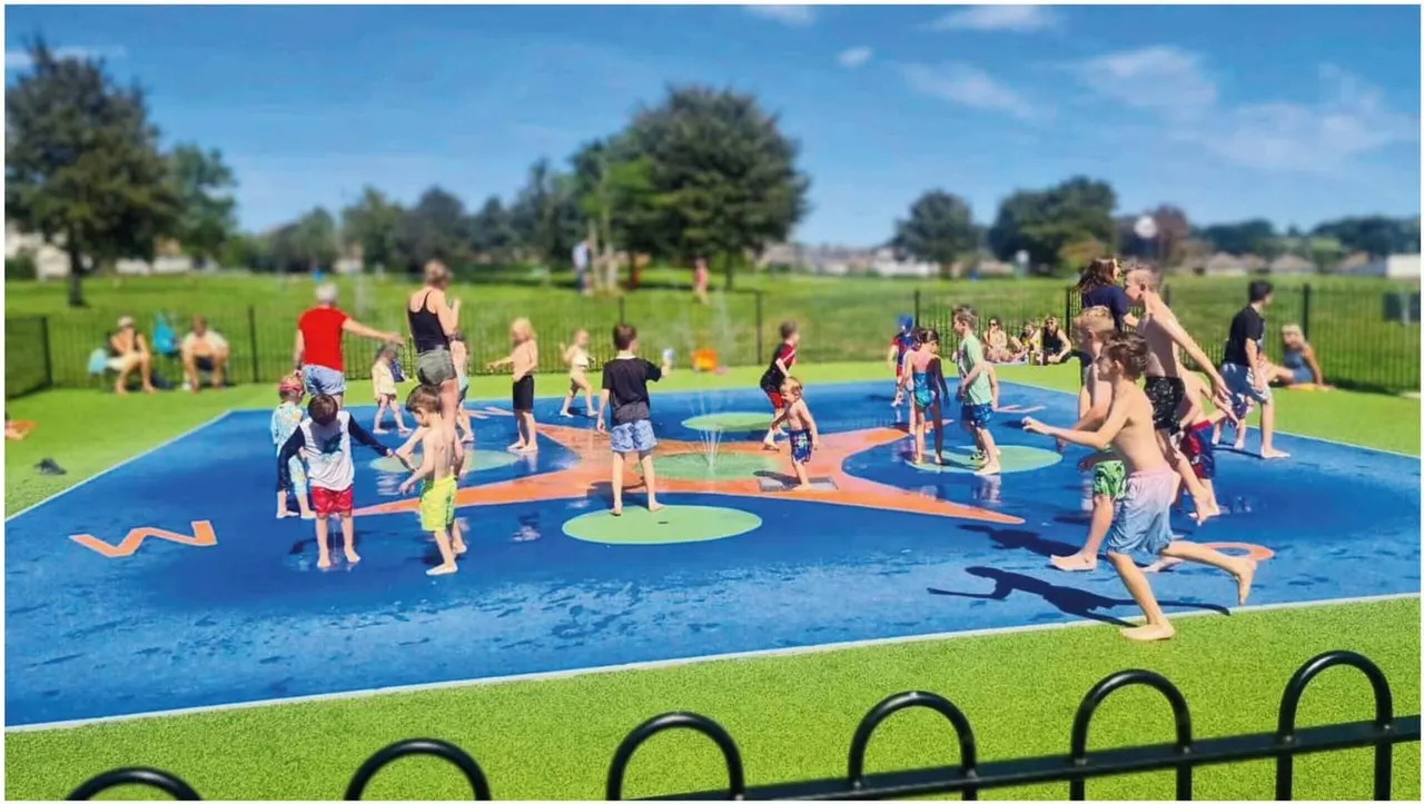 Children and adults playing and splashing in a shallow blue water pad surrounded by green grass and trees on a sunny day.