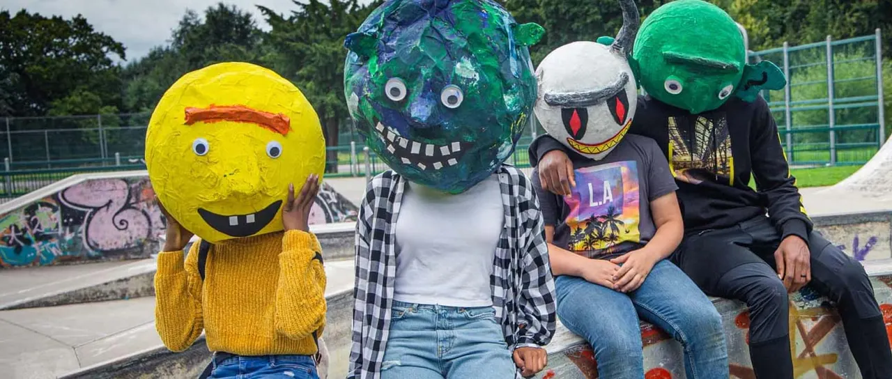 Four people wearing large painted ball heads pose together at a skate park with ramps and graffiti in the background.