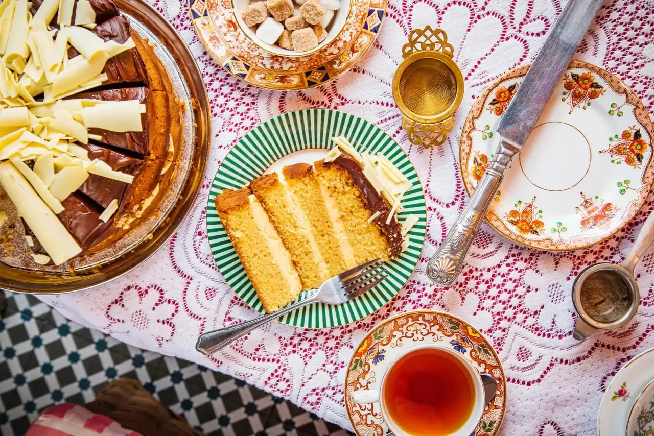 A slice of layered sponge cake with chocolate filling on a green-striped plate, surrounded by cheese, sugar cubes, tea, and ornate tableware on a patterned tablecloth.