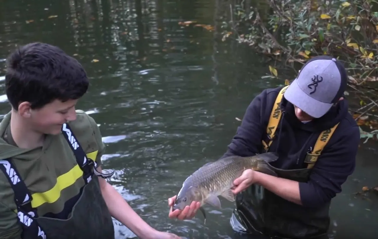 Two young people in waders examine a freshwater fish caught at the water's edge.