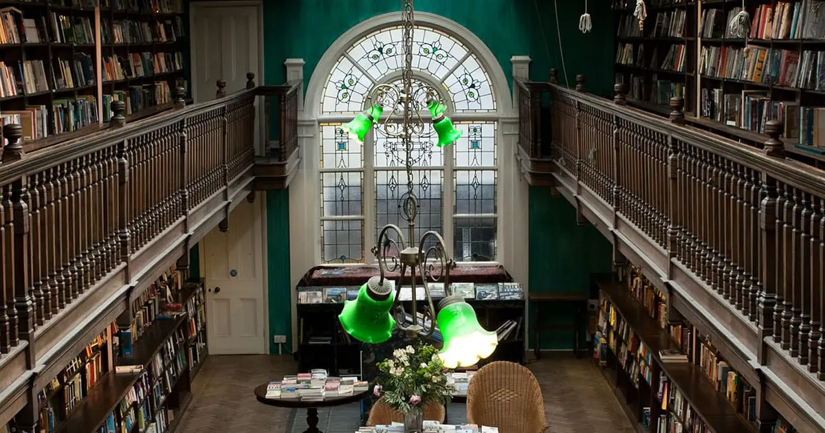 An elegant library interior with two-storey wooden bookshelves on either side, a large arched stained-glass window, and bright green pendant lamps hanging above a reading area with a plant and chair.