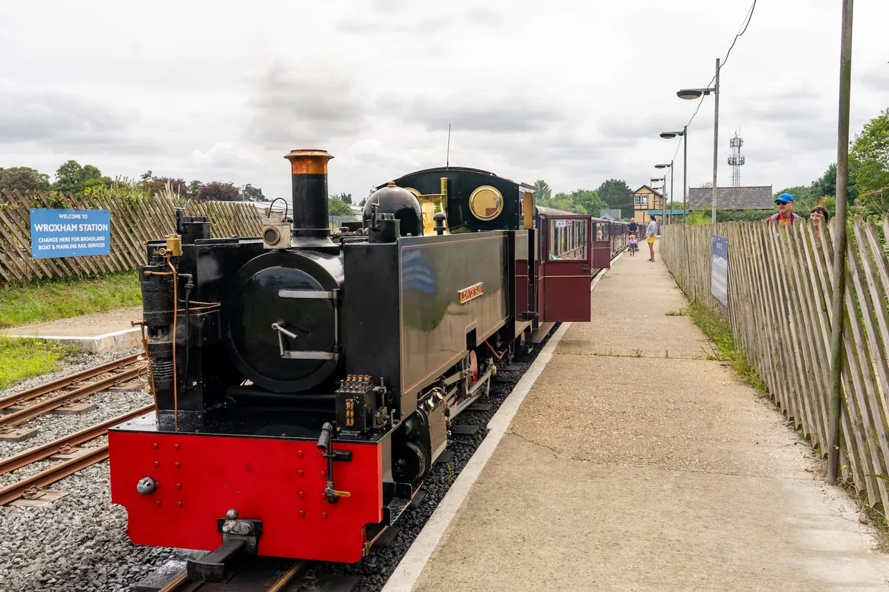 Heritage steam train with red and black locomotive at platform with passengers boarding maroon carriages.