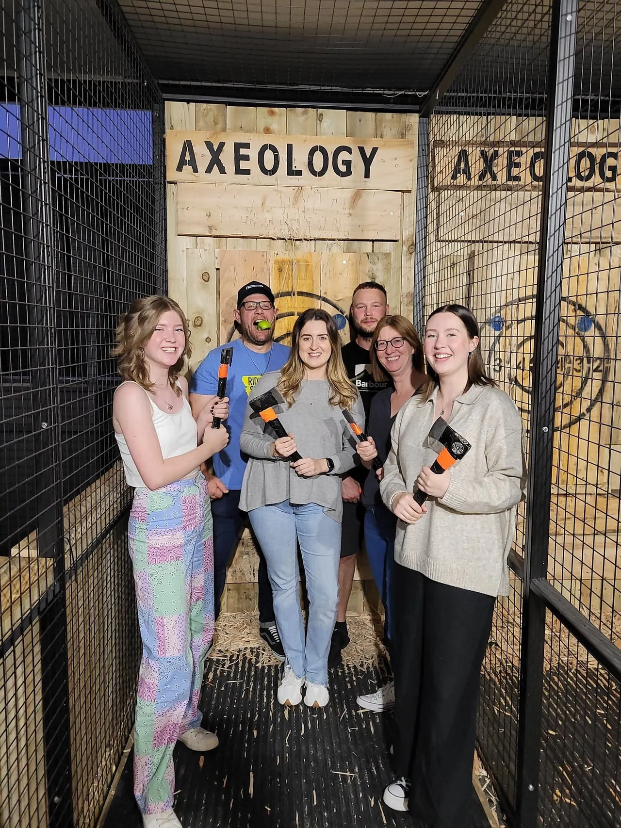 A group of six adults holding axes inside an axe-throwing venue with a wooden target wall and "AXEOLOGY" signage.