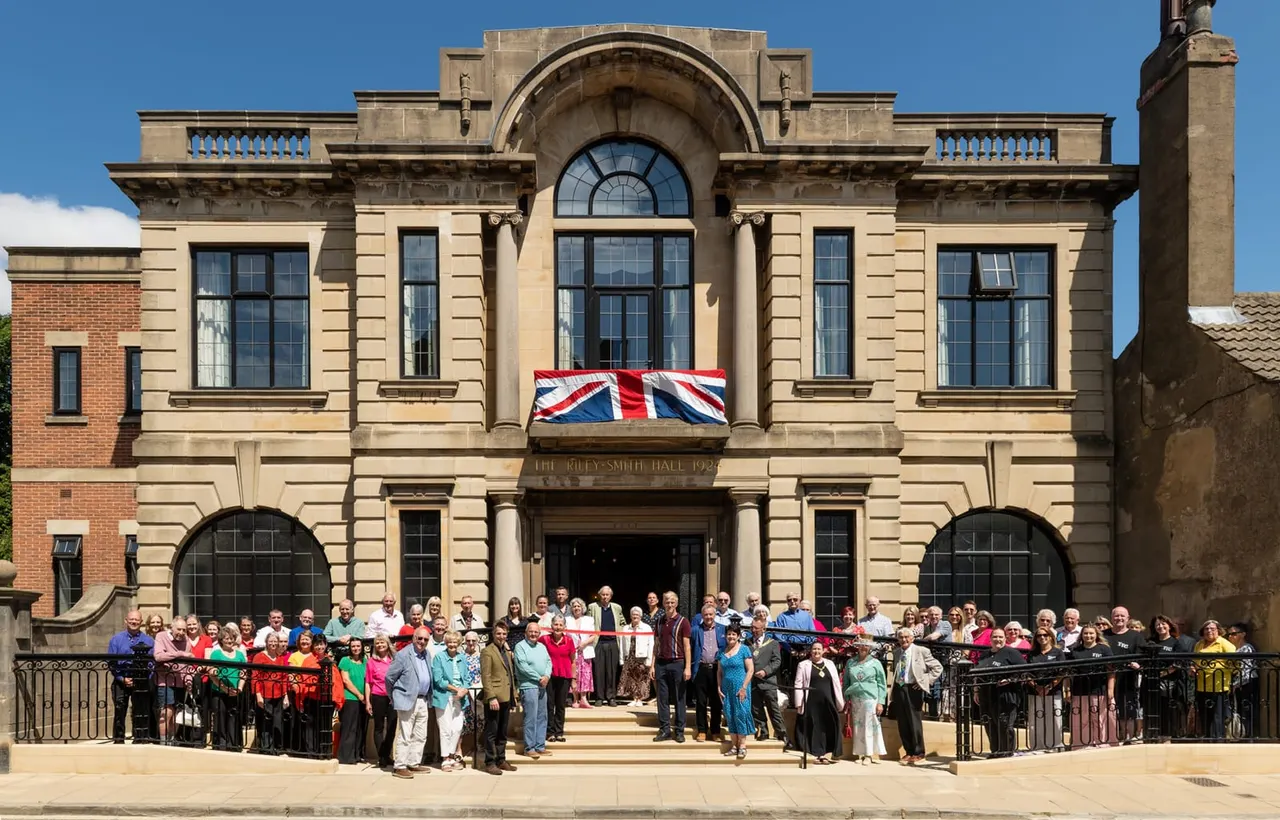 A large crowd of visitors gathered in front of a classical stone building with a Union Jack flag displayed above the entrance on a sunny day.