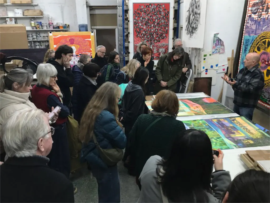 A group of visitors gathered in an artist's studio observing a presentation about colourful printed artworks displayed on tables and walls.