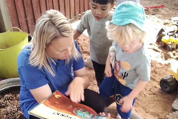 Woman and two young children looking at a book together in a sandy outdoor play area.