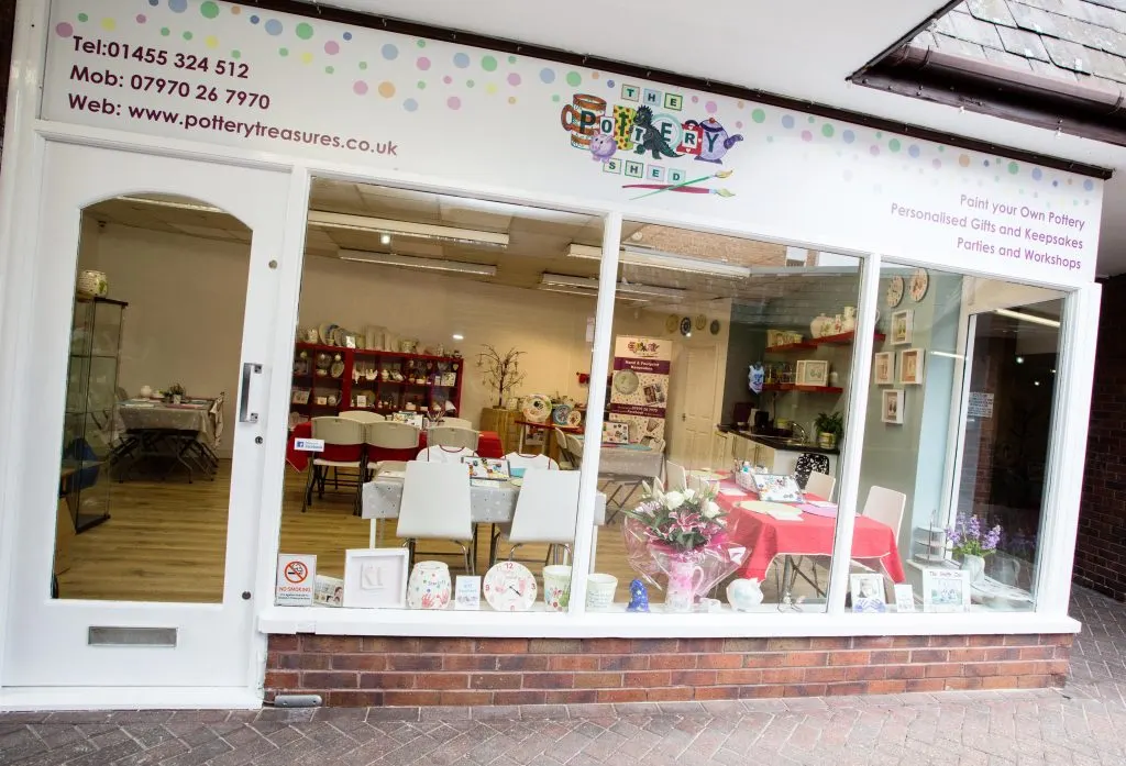 Shopfront window display for a pottery painting venue showing tables with white chairs, shelves of ceramic pieces, and red decorative accents inside.