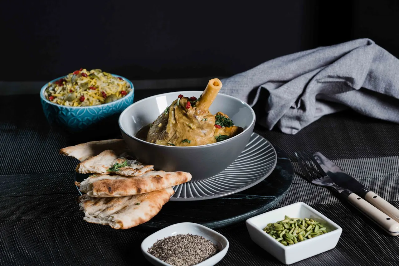 Plated Persian meal with roasted meat in sauce, grilled fish fillets, rice dish, and side condiments on dark table.