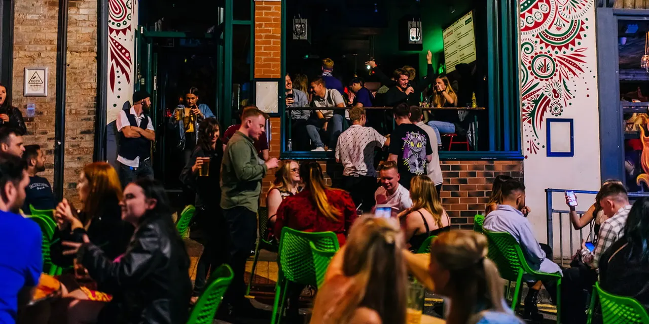 Busy outdoor bar terrace at night with guests on green chairs drinking and socialising in front of an illuminated brick venue.