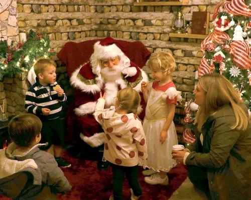 Father Christmas in red suit and white beard distributes gifts to a group of young children by a brick fireplace decorated with Christmas garlands.