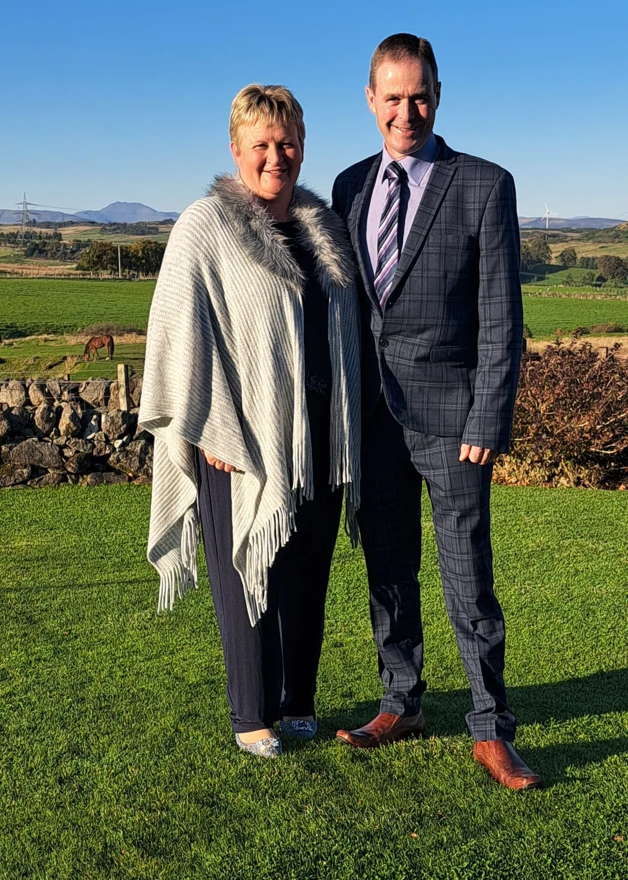 Two people standing on a grass field with rural farmland, stone walls and mountains visible behind them.