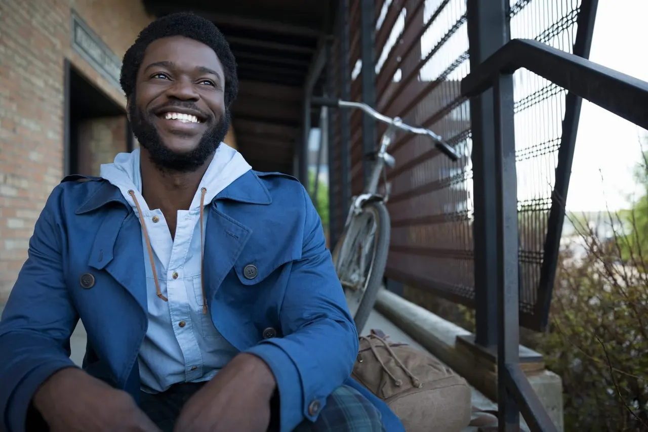 Man in blue jacket smiling at camera whilst sitting on steps outside a brick building with metal fencing.