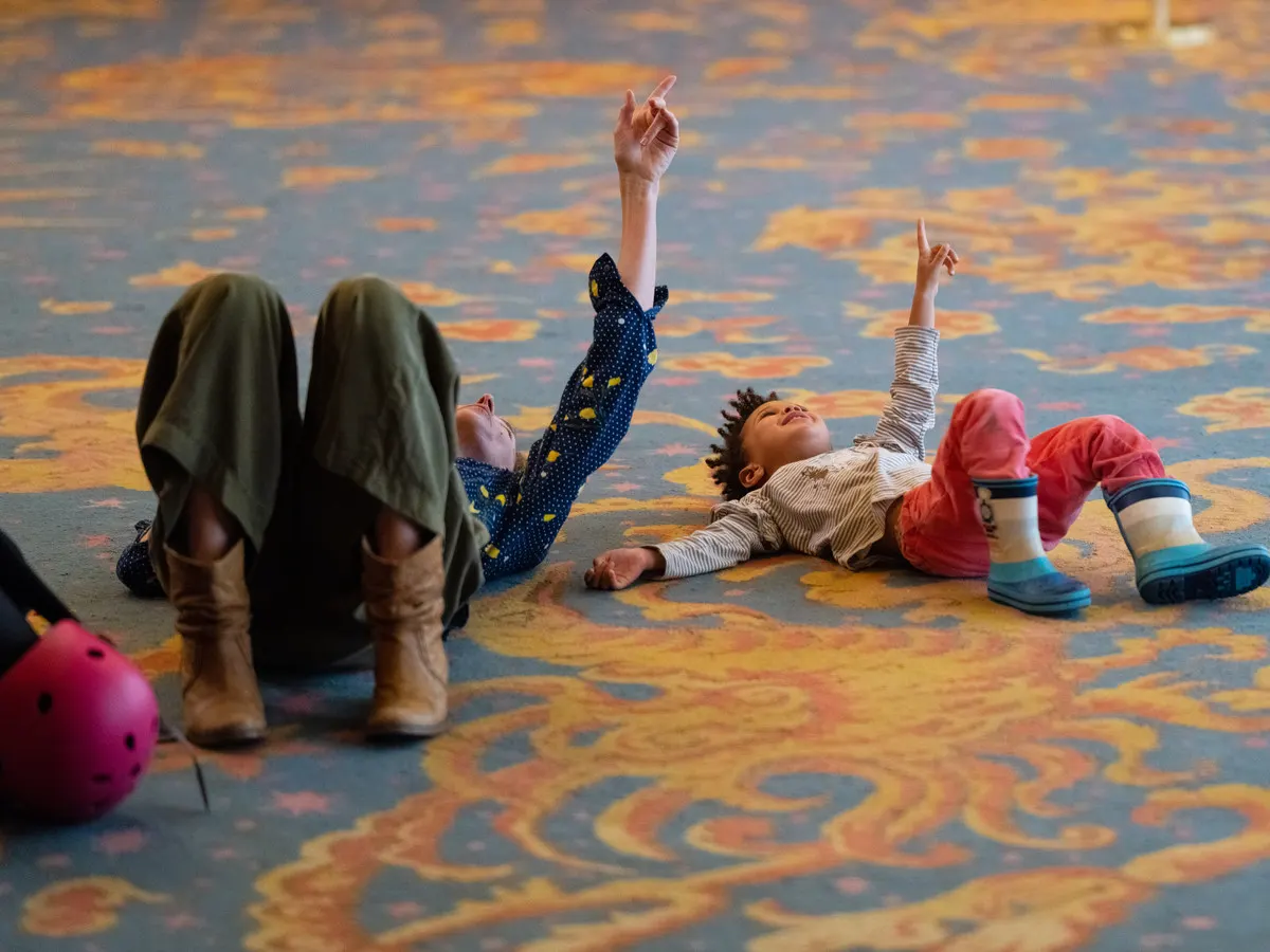 Young children playing and lying on a colourful patterned floor indoors, stretching their arms upwards.