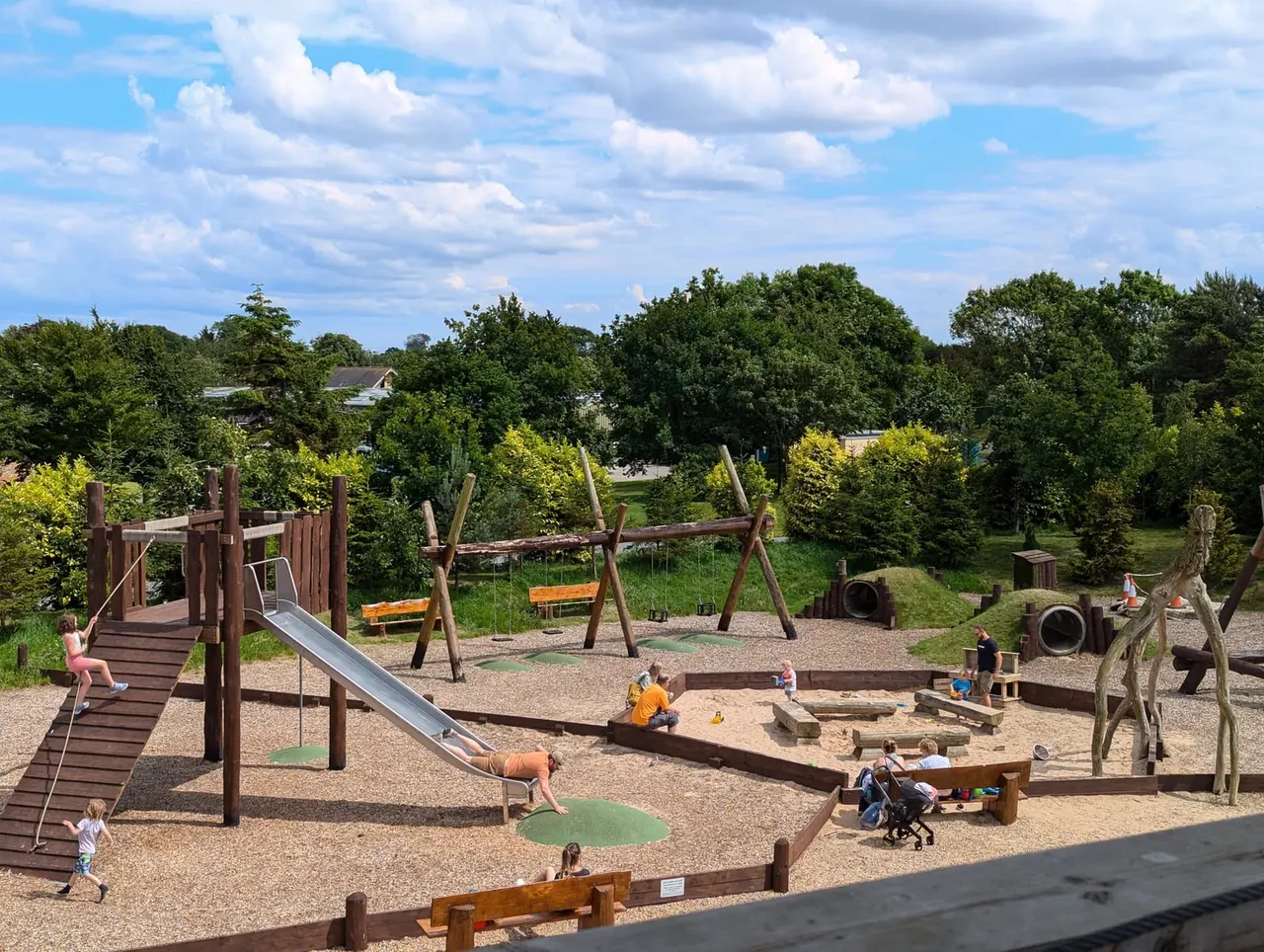 Children playing in a natural adventure playground with wooden climbing structures, slides, swings and sand areas surrounded by trees.