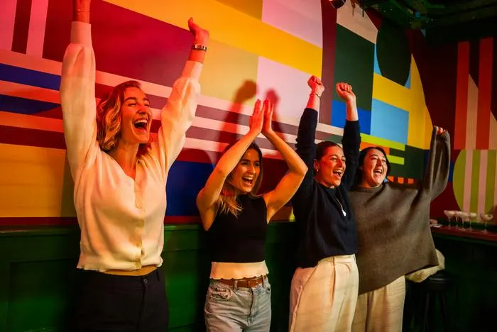 Four people celebrating with arms raised in front of a colourful striped wall at an indoor entertainment venue.