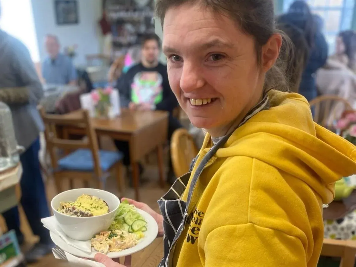 A woman in a yellow jacket holds a plate of food whilst smiling at a busy cafe interior with other visitors.
