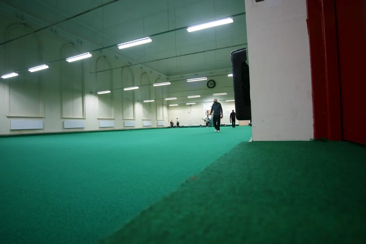 People walking on a green indoor bowling green with fluorescent lighting overhead.