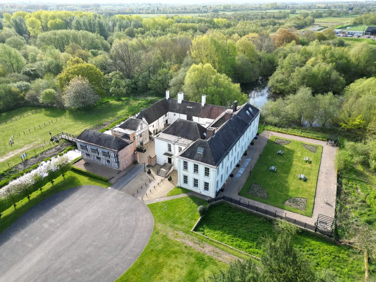 Aerial view of a historic white manor house with dark roofs set in landscaped grounds surrounded by woodland and a river.
