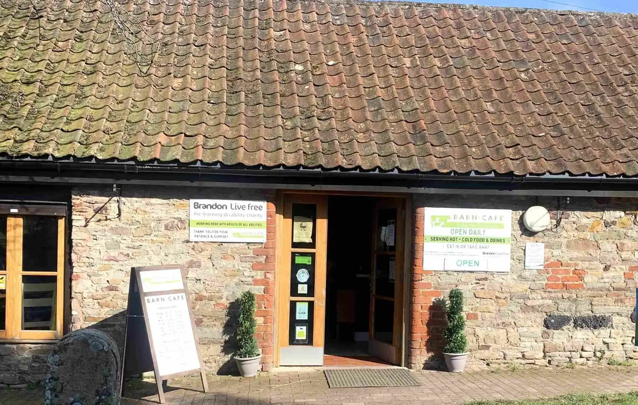 Stone barn café with wooden doors and windows, terracotta tile roof, open signage and potted plants flanking the entrance.