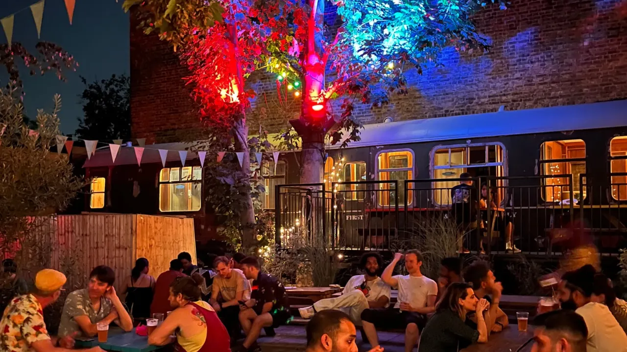 Evening outdoor event with guests seated at tables beneath colourful illuminated foliage and bunting, beside a converted train carriage venue.