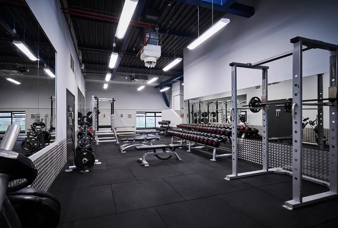 Modern gym interior with weight racks, dumbbells, benches and exercise equipment under bright lighting.