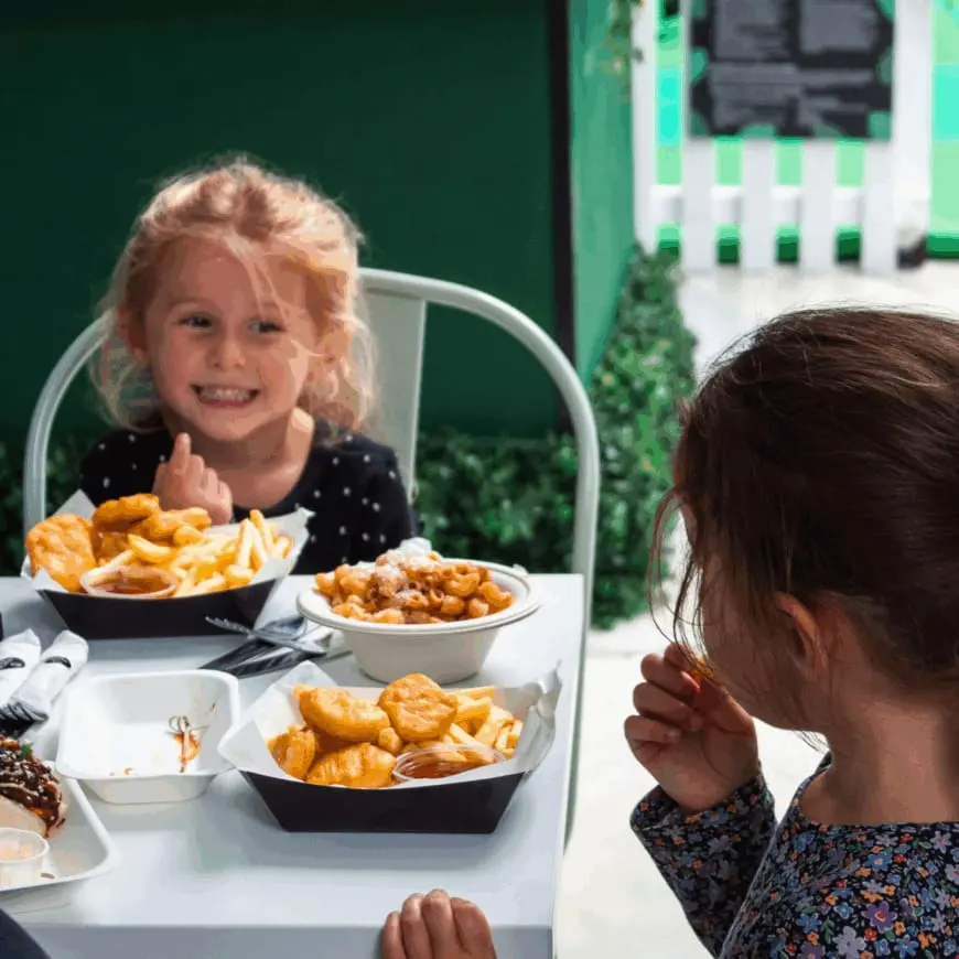 Child eating chips and fried food at an outdoor dining table, reflected in a mirror.