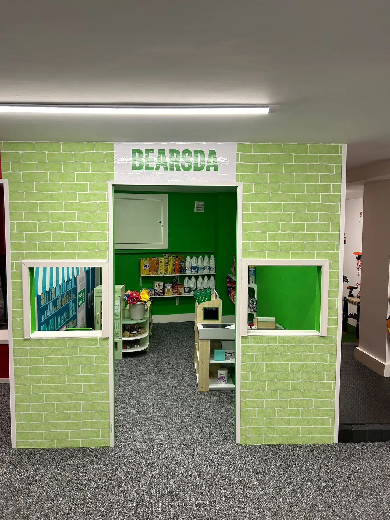 A green and white themed play structure with "BEARODA" signage, featuring shelves stocked with supplies and play equipment visible inside the entrance.